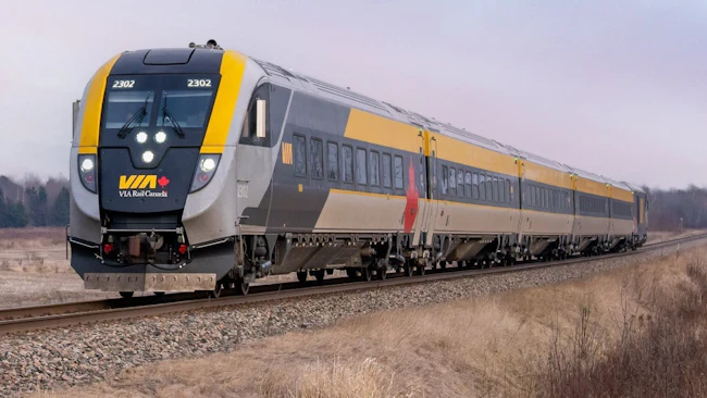 VIA Rail Canada Siemens Venture trainset number 2302 in the agency's gray and yellow livery with a Canadian maple leaf accent, traveling along a rural track through a winter landscape of dormant brush and overcast skies.