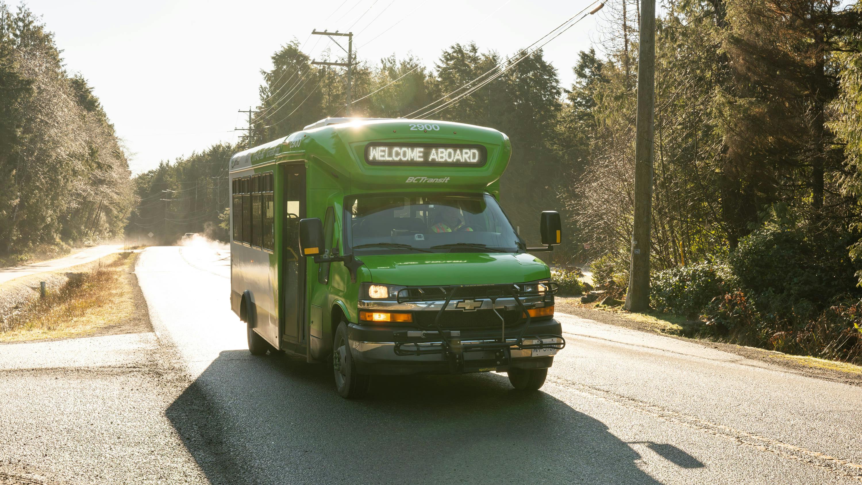 BC Transit small green Chevrolet cutaway bus number 2900 displaying 'Welcome Aboard' on its destination board travels along a narrow, sunlit rural road lined with evergreen trees in British Columbia.