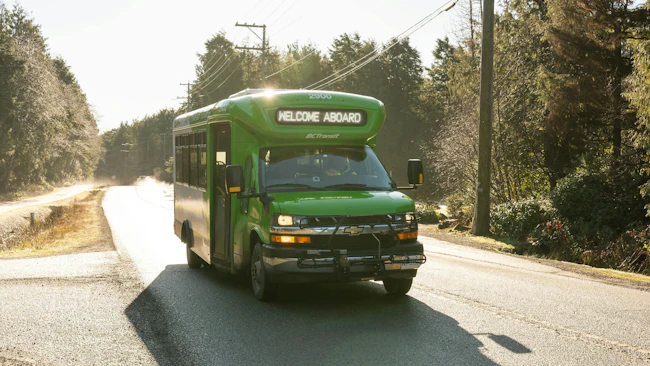 BC Transit small green Chevrolet cutaway bus number 2900 displaying 'Welcome Aboard' on its destination board travels along a narrow, sunlit rural road lined with evergreen trees in British Columbia.