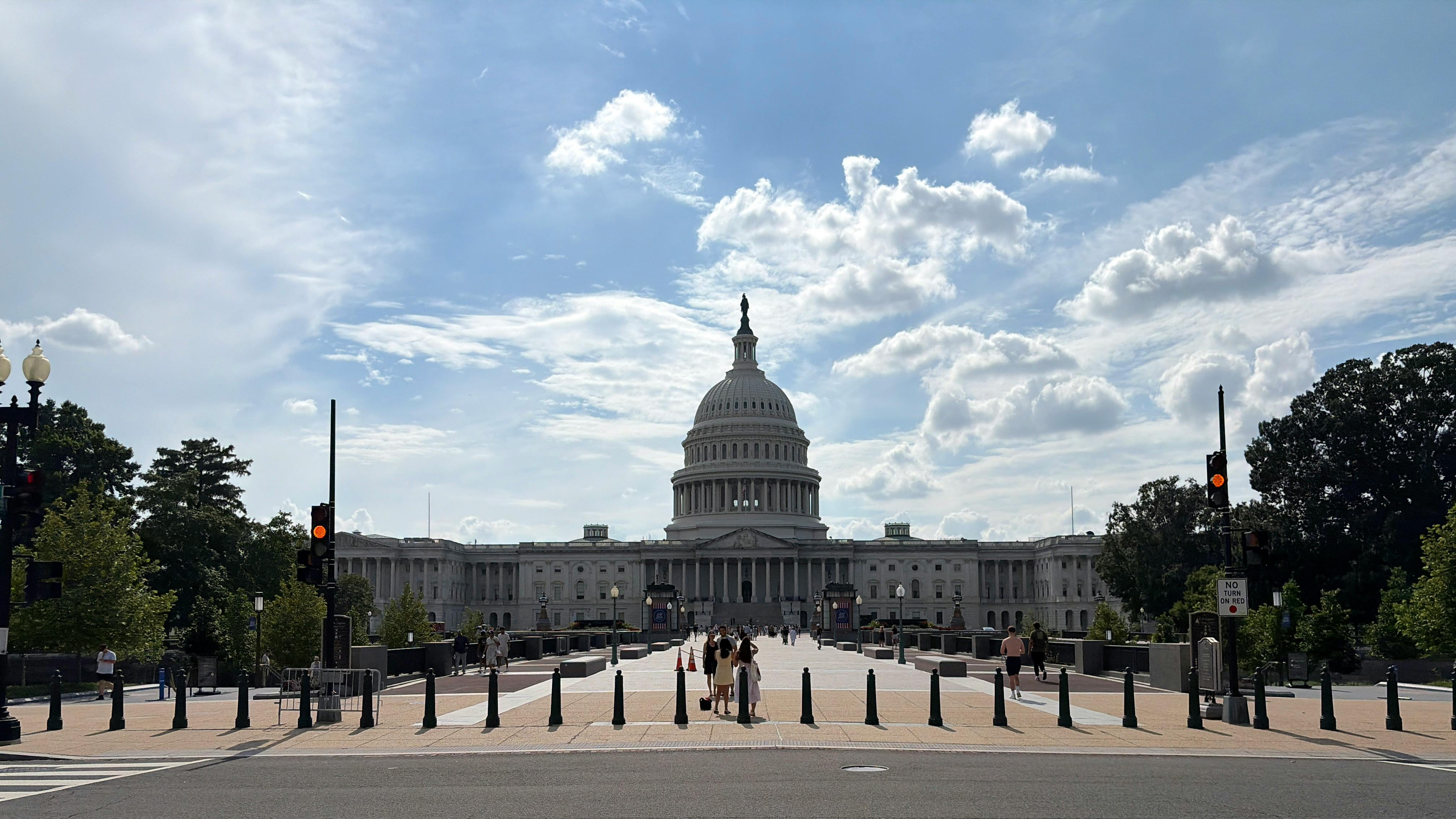 The U.S. Capitol Building's west front viewed from a bollard-lined pedestrian approach, with the central dome topped by the Statue of Freedom framed against a partly cloudy sky and a few visitors walking on the plaza.