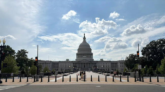 The U.S. Capitol Building's west front viewed from a bollard-lined pedestrian approach, with the central dome topped by the Statue of Freedom framed against a partly cloudy sky and a few visitors walking on the plaza.