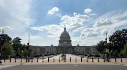 The U.S. Capitol Building's west front viewed from a bollard-lined pedestrian approach, with the central dome topped by the Statue of Freedom framed against a partly cloudy sky and a few visitors walking on the plaza.