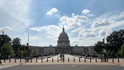 The U.S. Capitol Building's west front viewed from a bollard-lined pedestrian approach, with the central dome topped by the Statue of Freedom framed against a partly cloudy sky and a few visitors walking on the plaza.