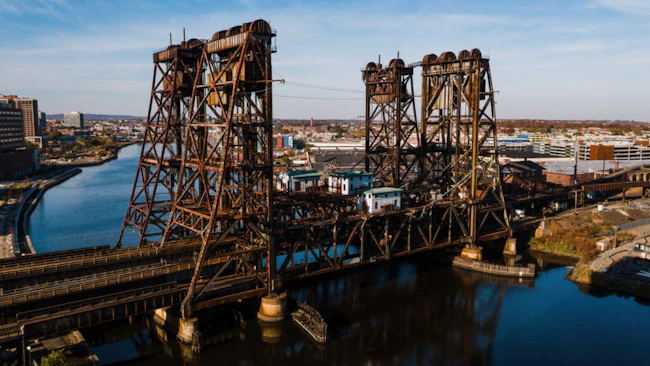 An aerial view of the aging Dock Bridge, a vertical lift railroad bridge spanning the Passaic River in Newark, New Jersey, featuring two large rusted steel lift towers and a movable span with a small operator's house, with the Newark skyline visible in the background under a clear autumn sky.