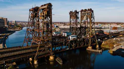 An aerial view of the aging Dock Bridge, a vertical lift railroad bridge spanning the Passaic River in Newark, New Jersey, featuring two large rusted steel lift towers and a movable span with a small operator's house, with the Newark skyline visible in the background under a clear autumn sky.