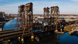 An aerial view of the aging Dock Bridge, a vertical lift railroad bridge spanning the Passaic River in Newark, New Jersey, featuring two large rusted steel lift towers and a movable span with a small operator's house, with the Newark skyline visible in the background under a clear autumn sky.