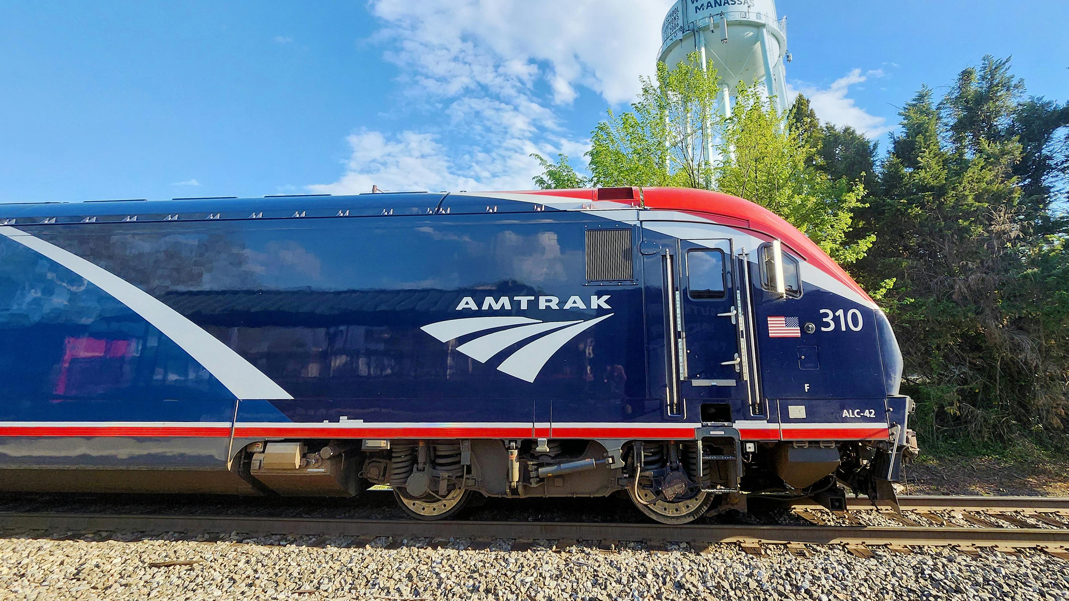 A blue, white and red Amtrak ALC-42 Charger locomotive, numbered 310 and bearing the agency's wordmark and arrow logo, sits on track under a partly cloudy blue sky with trees in the background.