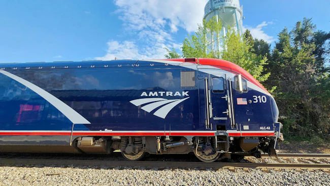 A blue, white and red Amtrak ALC-42 Charger locomotive, numbered 310 and bearing the agency's wordmark and arrow logo, sits on track under a partly cloudy blue sky with trees in the background.