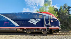 A blue, white and red Amtrak ALC-42 Charger locomotive, numbered 310 and bearing the agency's wordmark and arrow logo, sits on track under a partly cloudy blue sky with trees in the background.