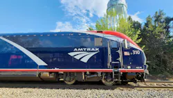 A blue, white and red Amtrak ALC-42 Charger locomotive, numbered 310 and bearing the agency's wordmark and arrow logo, sits on track under a partly cloudy blue sky with trees in the background.