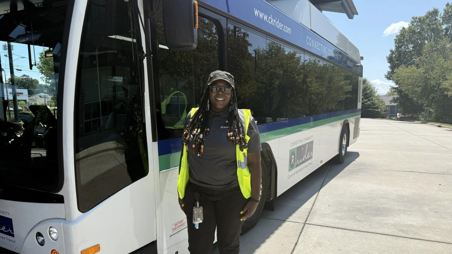 WeDriveU bus operator Charlene Miller stands smiling in a yellow safety vest beside a Concord Kannapolis Area Transit (CK Rider) electric bus displaying the agency's 'Connecting Community' branding and ckrider.com website on a sunny day.