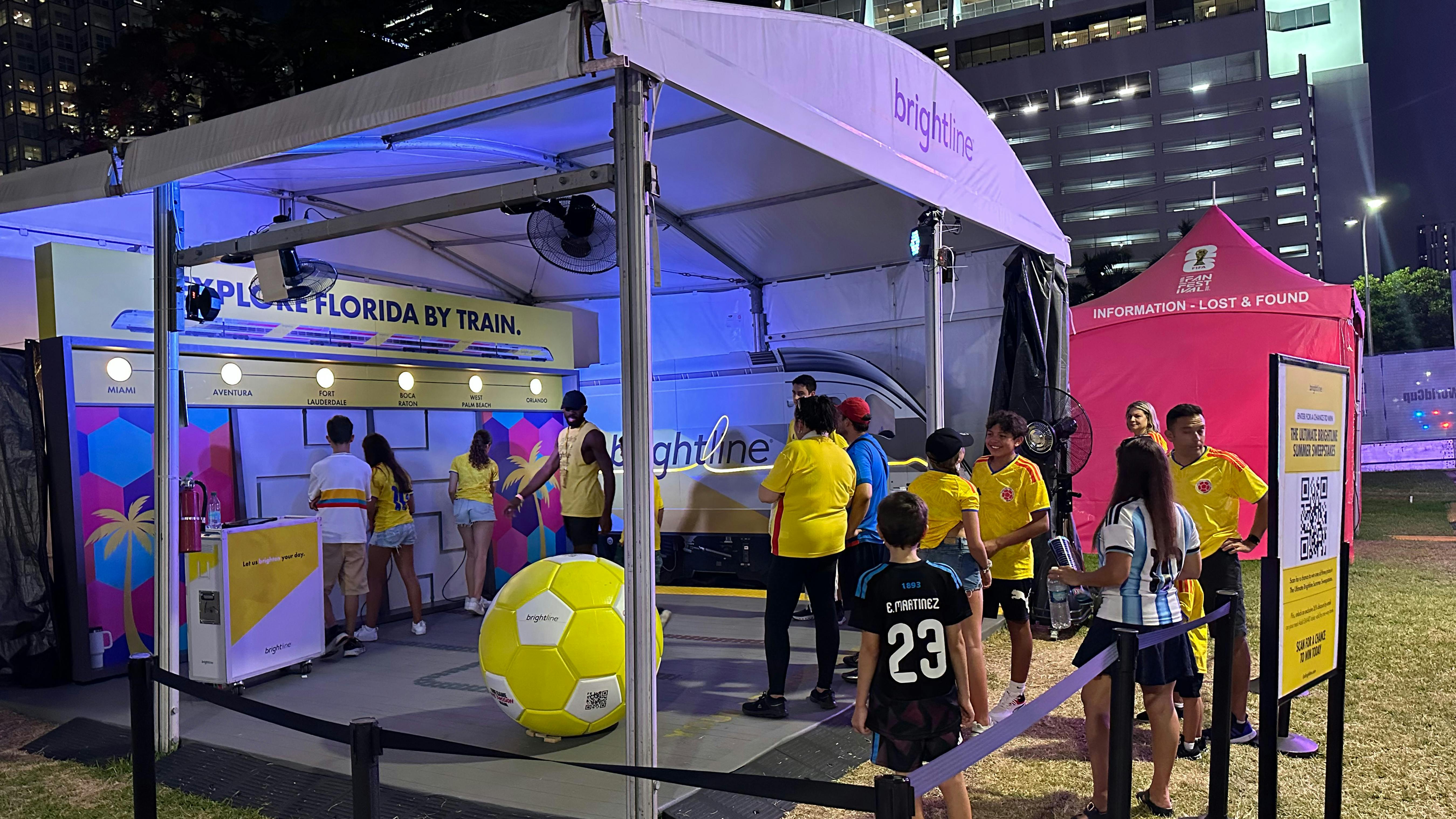 A Brightline branded experiential marketing tent at a FIFA World Cup 2026 fan event in Miami, featuring an 'Explore Florida by Train' display listing stations from Miami to Orlando, a large yellow Brightline-branded soccer ball, and soccer fans in national team jerseys engaging with staff at night.
