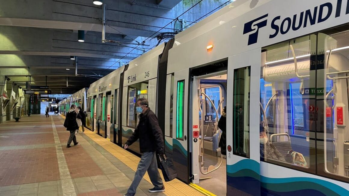 Passengers board and walk alongside Sound Transit Link light rail car 246 at an underground station platform, with the train's doors open and its white, navy, and teal wave livery visible, and the illuminated interior showing seated riders