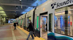 Passengers board and walk alongside Sound Transit Link light rail car 246 at an underground station platform, with the train's doors open and its white, navy, and teal wave livery visible, and the illuminated interior showing seated riders