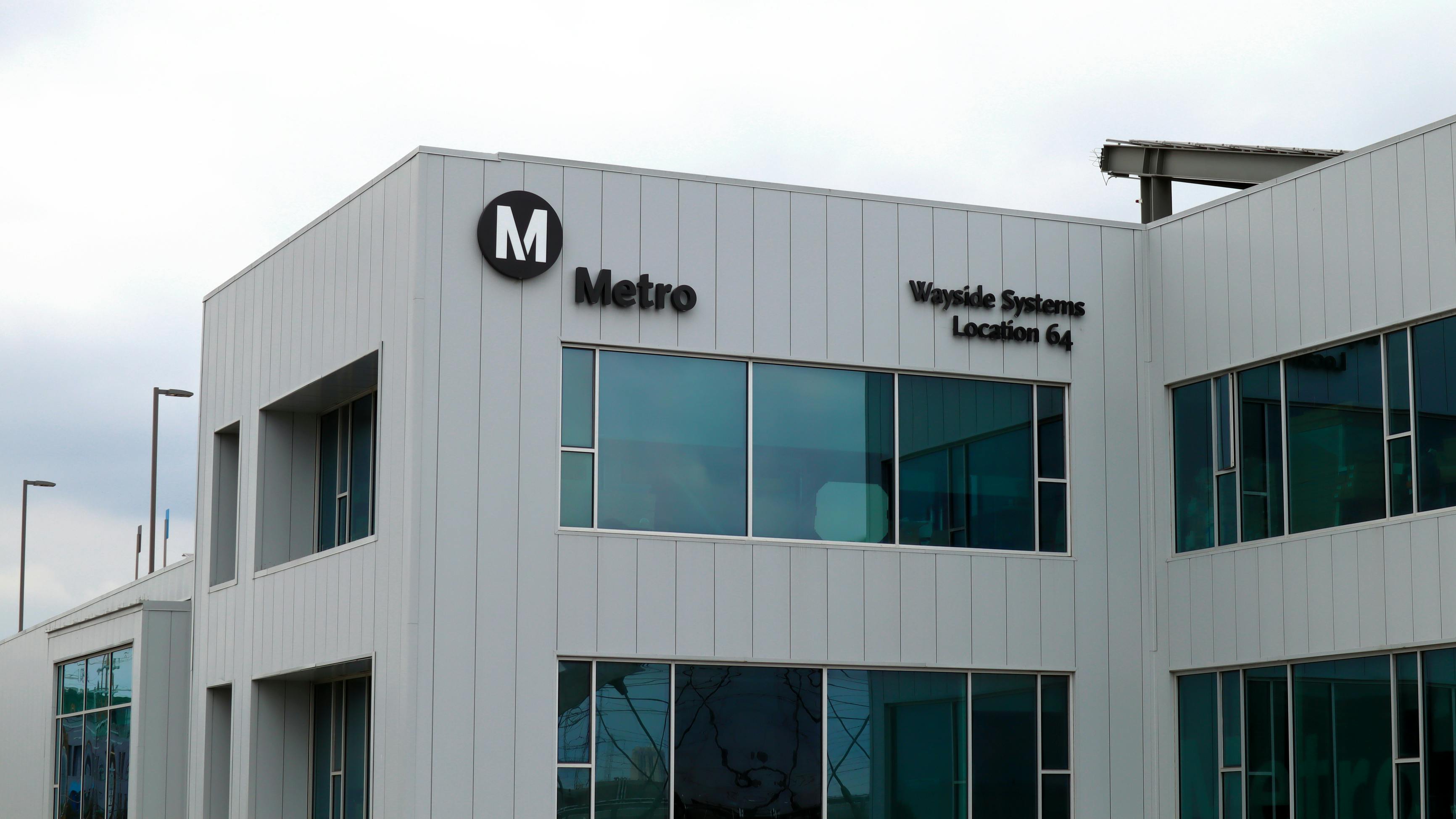 The exterior of Los Angeles Metro's Wayside Systems Location 64 facility, a modern two-story building with vertical metal siding and large windows, marked with the agency's black circular 'M' Metro logo and facility name.