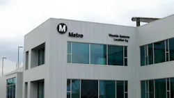 The exterior of Los Angeles Metro's Wayside Systems Location 64 facility, a modern two-story building with vertical metal siding and large windows, marked with the agency's black circular 'M' Metro logo and facility name.