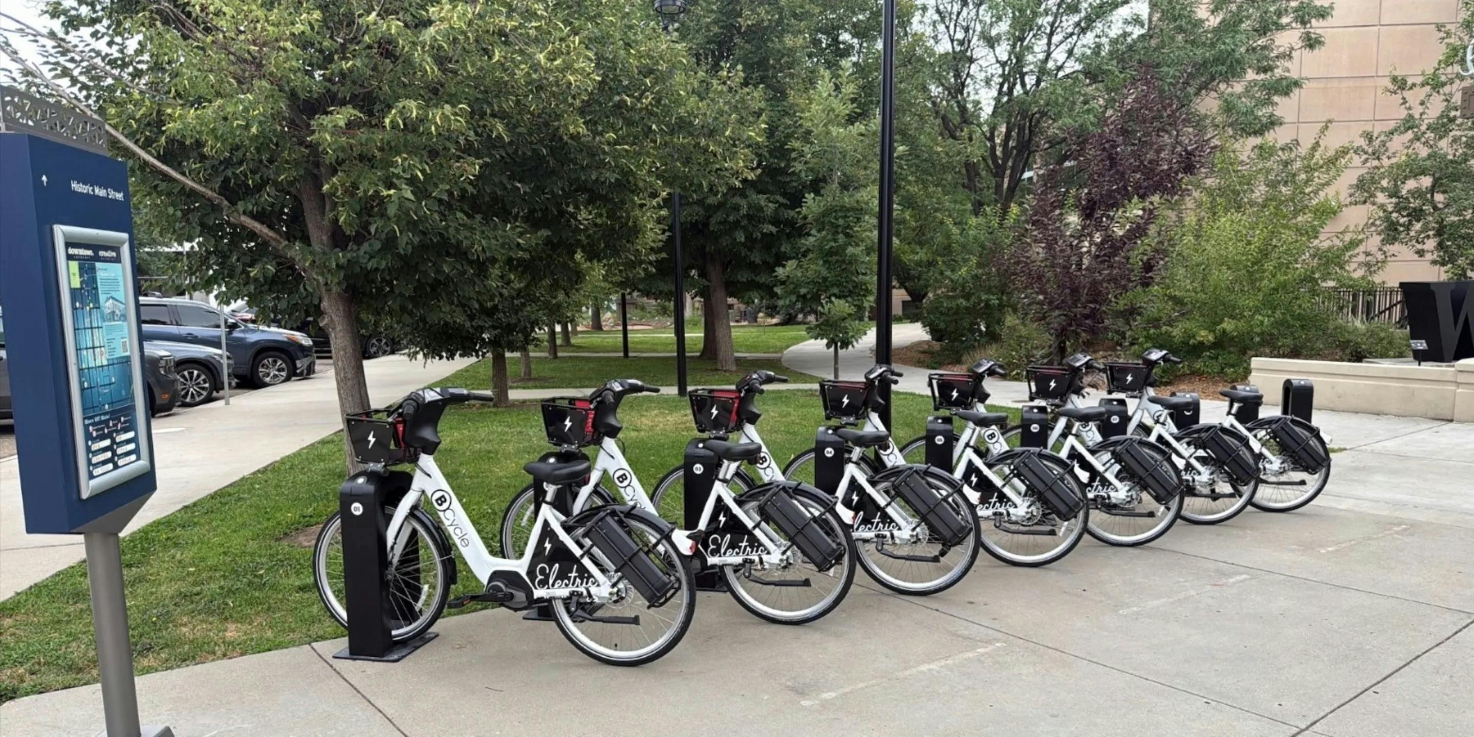 A row of white and black BCycle electric bikeshare bicycles docked at a station along Historic Main Street, with a wayfinding kiosk at left and mature trees and green space in the background.