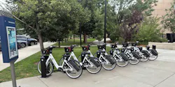A row of white and black BCycle electric bikeshare bicycles docked at a station along Historic Main Street, with a wayfinding kiosk at left and mature trees and green space in the background.