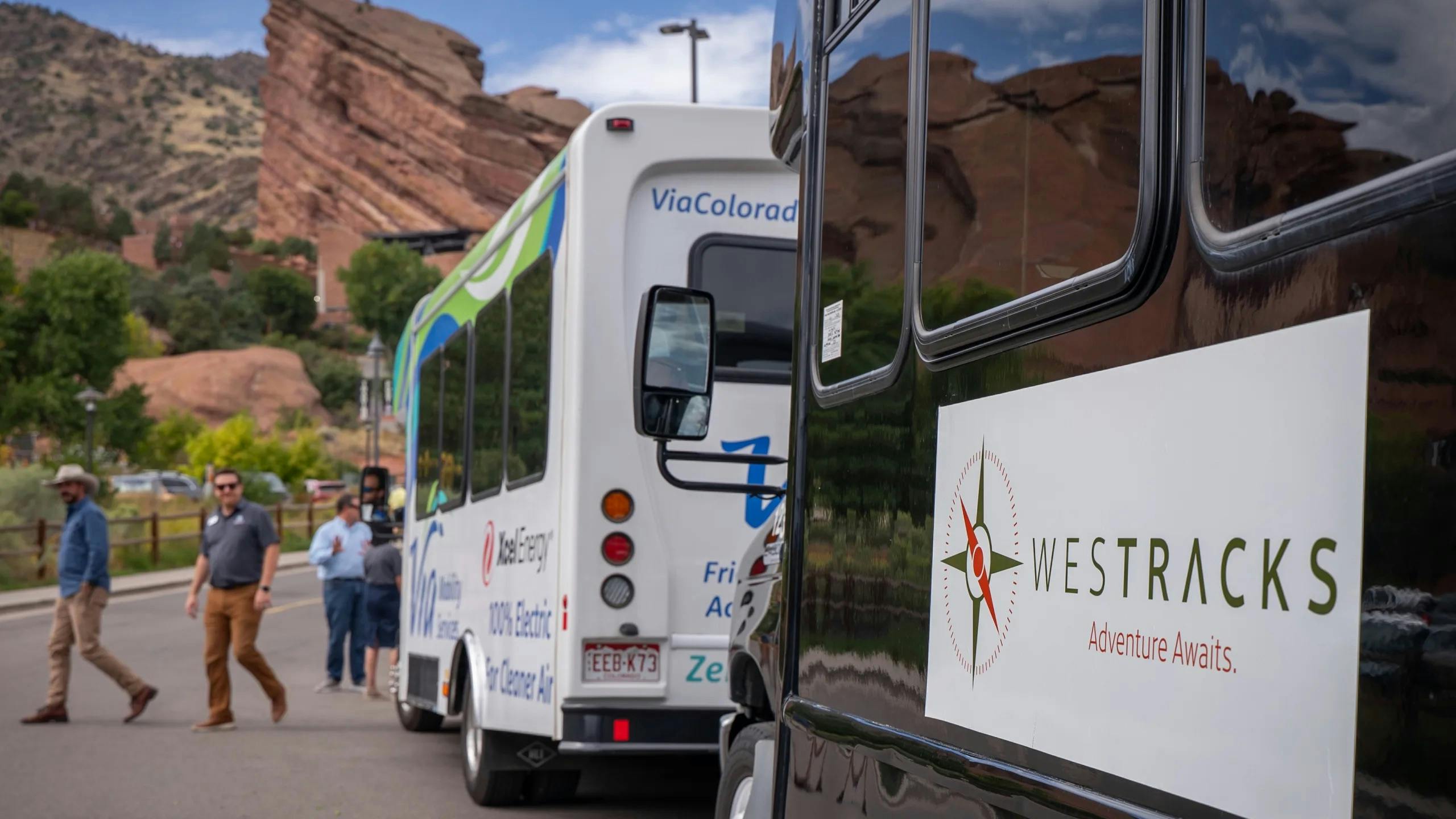A black WesTracks shuttle marked 'Adventure Awaits' is parked in the foreground with a white Via Colorado 100% electric shuttle in the background, alongside Red Rocks Park's distinctive red sandstone formations, as several passengers walk toward the vehicles.