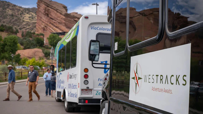 A black WesTracks shuttle marked 'Adventure Awaits' is parked in the foreground with a white Via Colorado 100% electric shuttle in the background, alongside Red Rocks Park's distinctive red sandstone formations, as several passengers walk toward the vehicles.
