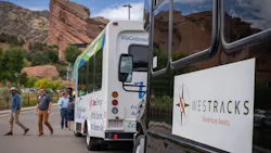 A black WesTracks shuttle marked 'Adventure Awaits' is parked in the foreground with a white Via Colorado 100% electric shuttle in the background, alongside Red Rocks Park's distinctive red sandstone formations, as several passengers walk toward the vehicles.