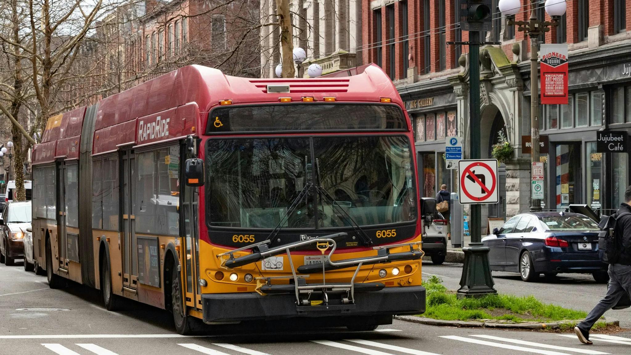 King County Metro RapidRide articulated bus number 6055 in the signature red and yellow livery navigates an intersection in Seattle's Pioneer Square neighborhood, with historic brick storefronts, a pedestrian crossing, and a no-left-turn sign visible in the background.