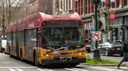King County Metro RapidRide articulated bus number 6055 in the signature red and yellow livery navigates an intersection in Seattle's Pioneer Square neighborhood, with historic brick storefronts, a pedestrian crossing, and a no-left-turn sign visible in the background.