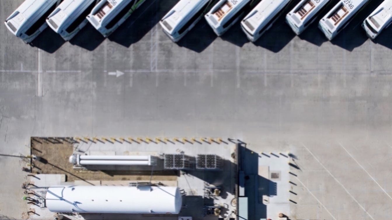 An overhead drone view of a Foothill Transit bus yard showing a row of parked white and green buses angled diagonally along a concrete apron, with a hydrogen fueling station and storage tanks visible below.