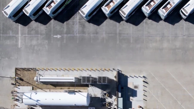 An overhead drone view of a Foothill Transit bus yard showing a row of parked white and green buses angled diagonally along a concrete apron, with a hydrogen fueling station and storage tanks visible below.