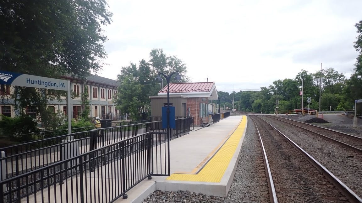 The newly renovated Amtrak platform at Huntingdon, Pa., showing a concrete platform with yellow tactile edge strips, a small brick shelter with a red metal roof, black railings and station signage, with a historic multi-story brick building visible to the left and the tracks curving off into the distance.