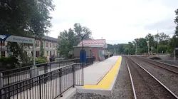 The newly renovated Amtrak platform at Huntingdon, Pa., showing a concrete platform with yellow tactile edge strips, a small brick shelter with a red metal roof, black railings and station signage, with a historic multi-story brick building visible to the left and the tracks curving off into the distance.