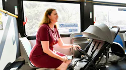 A woman in a red dress sits inside a New York City Transit bus with a folded stroller secured beside her in the newly designated stroller area, while a man in a suit stands nearby holding a yellow grab bar, with clear signage above explaining stroller securement rules.