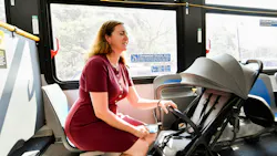 A woman in a red dress sits inside a New York City Transit bus with a folded stroller secured beside her in the newly designated stroller area, while a man in a suit stands nearby holding a yellow grab bar, with clear signage above explaining stroller securement rules.