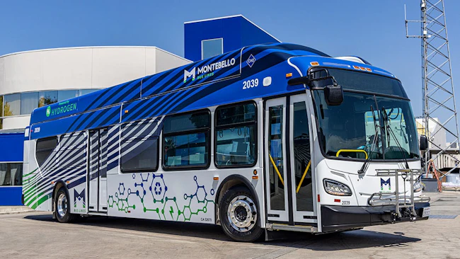 Montebello Bus Lines hydrogen fuel cell bus number 2039 in a bold blue, white, and gray wave livery with molecular chain graphics and a CH2 hydrogen fuel designation on the roof, parked in front of a hydrogen fueling facility on a clear day in Montebello, Calif.