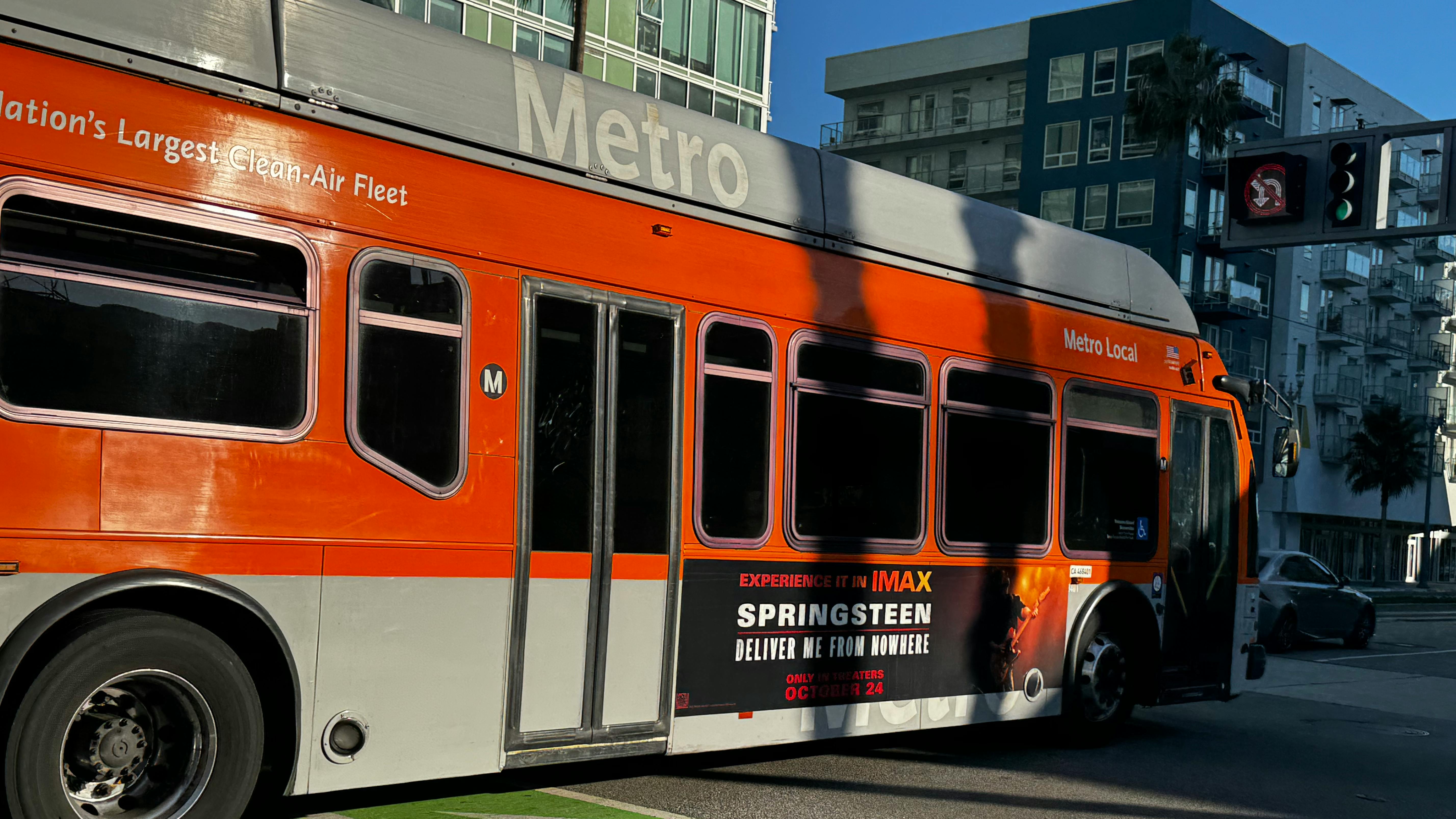 An LA Metro Local bus in the agency's orange and gray livery, bearing a side advertisement for the IMAX film 'Springsteen: Deliver Me from Nowhere,' passes through a Los Angeles intersection with a green-painted bike lane marking in the foreground and mid-rise residential buildings visible in the background.