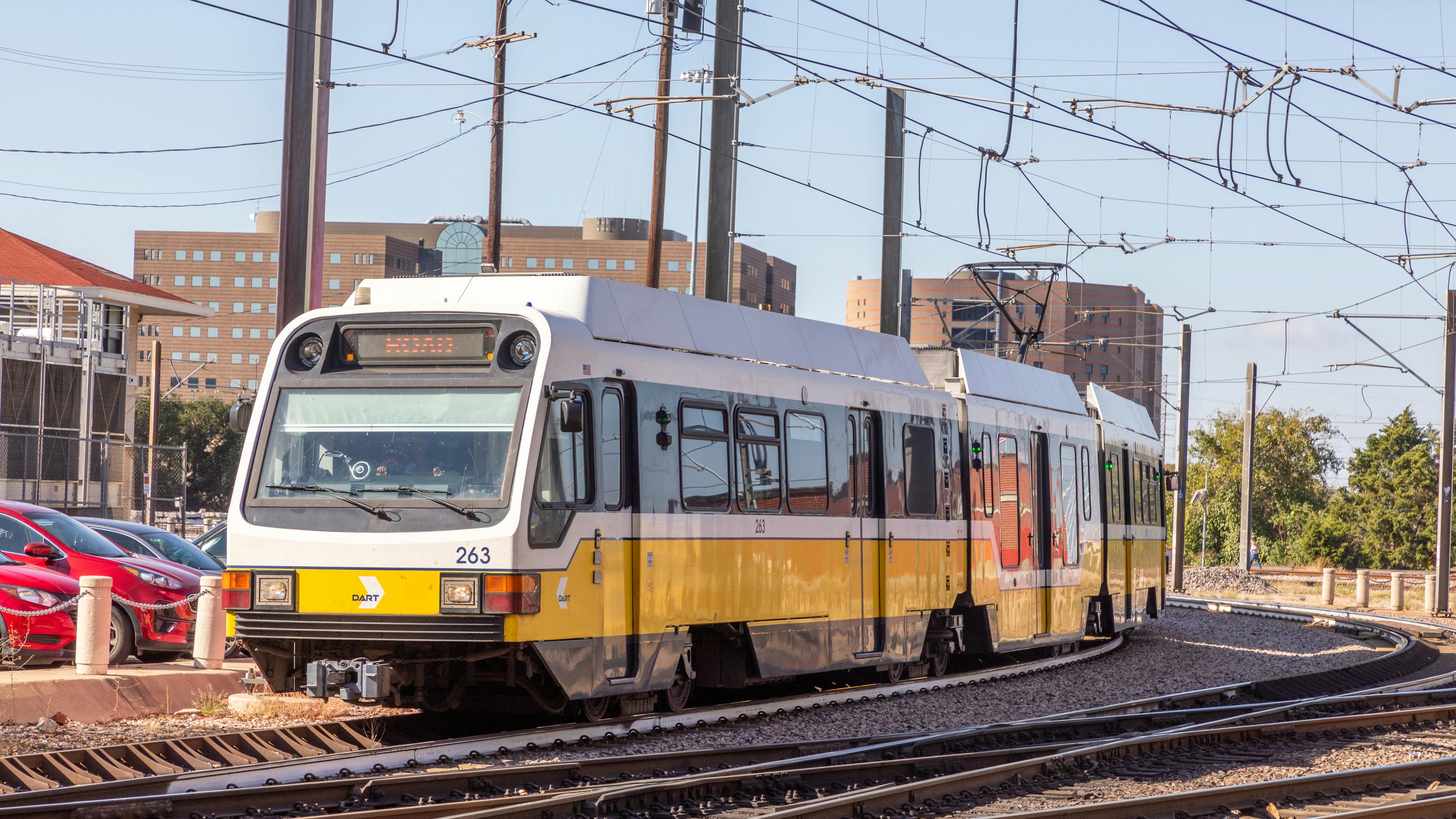 A DART light rail vehicle, numbered 263, rounds a curve on outdoor tracks near overhead catenary wires, with parked cars and mid-rise buildings visible in the background.