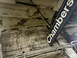 A close-up view of severe infrastructure deterioration at the MTA New York City Transit Chambers Street subway station, showing a heavily water-damaged and crumbling ceiling with peeling paint, exposed brick, staining, and corrosion alongside the station's black and white hanging platform sign.