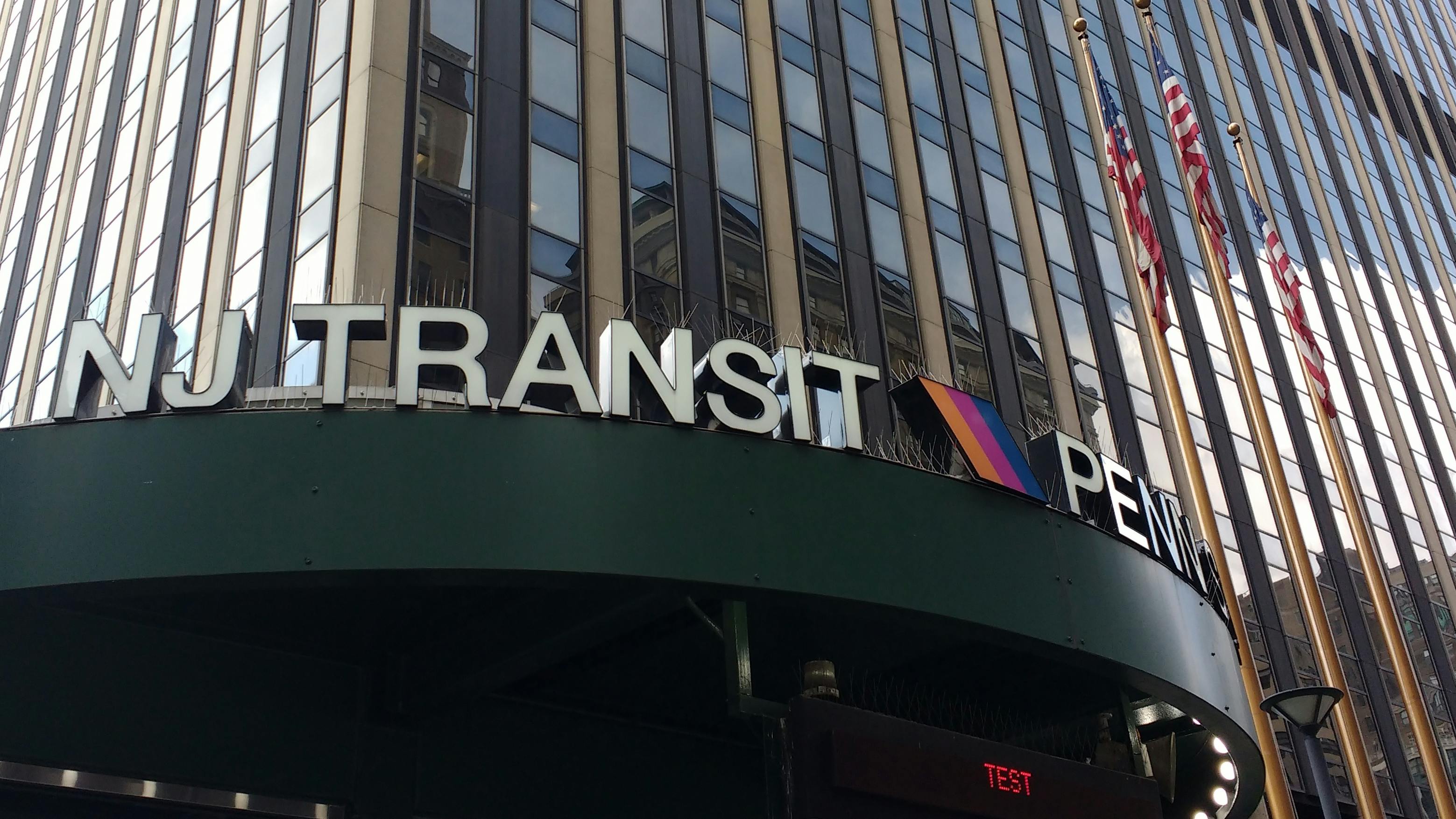 Close-up view of the NJ Transit sign at Penn Station's exterior entrance in New York City, with the agency's striped logo mounted on a dark green canopy below a tall office tower with vertical fa&ccedil;ade columns and American flags.