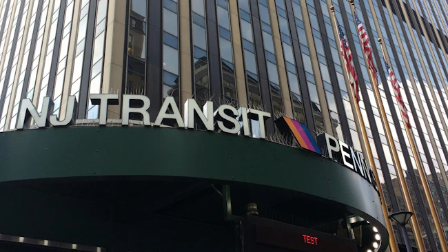 Close-up view of the NJ Transit sign at Penn Station's exterior entrance in New York City, with the agency's striped logo mounted on a dark green canopy below a tall office tower with vertical fa&ccedil;ade columns and American flags.