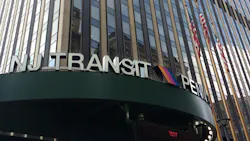 Close-up view of the NJ Transit sign at Penn Station's exterior entrance in New York City, with the agency's striped logo mounted on a dark green canopy below a tall office tower with vertical façade columns and American flags.
