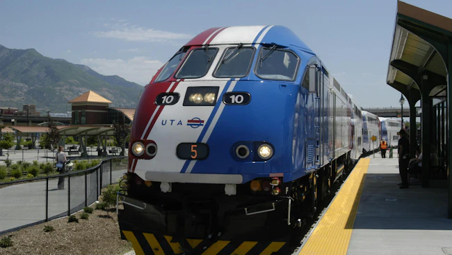 Utah Transit Authority FrontRunner commuter rail locomotive number 10 in a red, white and blue patriotic livery sits at a station platform with the Wasatch Mountains visible in the background on a clear sunny day, with passengers and a staff member in an orange safety vest visible along the platform.
