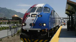 Utah Transit Authority FrontRunner commuter rail locomotive number 10 in a red, white and blue patriotic livery sits at a station platform with the Wasatch Mountains visible in the background on a clear sunny day, with passengers and a staff member in an orange safety vest visible along the platform.