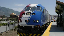 Utah Transit Authority FrontRunner commuter rail locomotive number 10 in a red, white and blue patriotic livery sits at a station platform with the Wasatch Mountains visible in the background on a clear sunny day, with passengers and a staff member in an orange safety vest visible along the platform.
