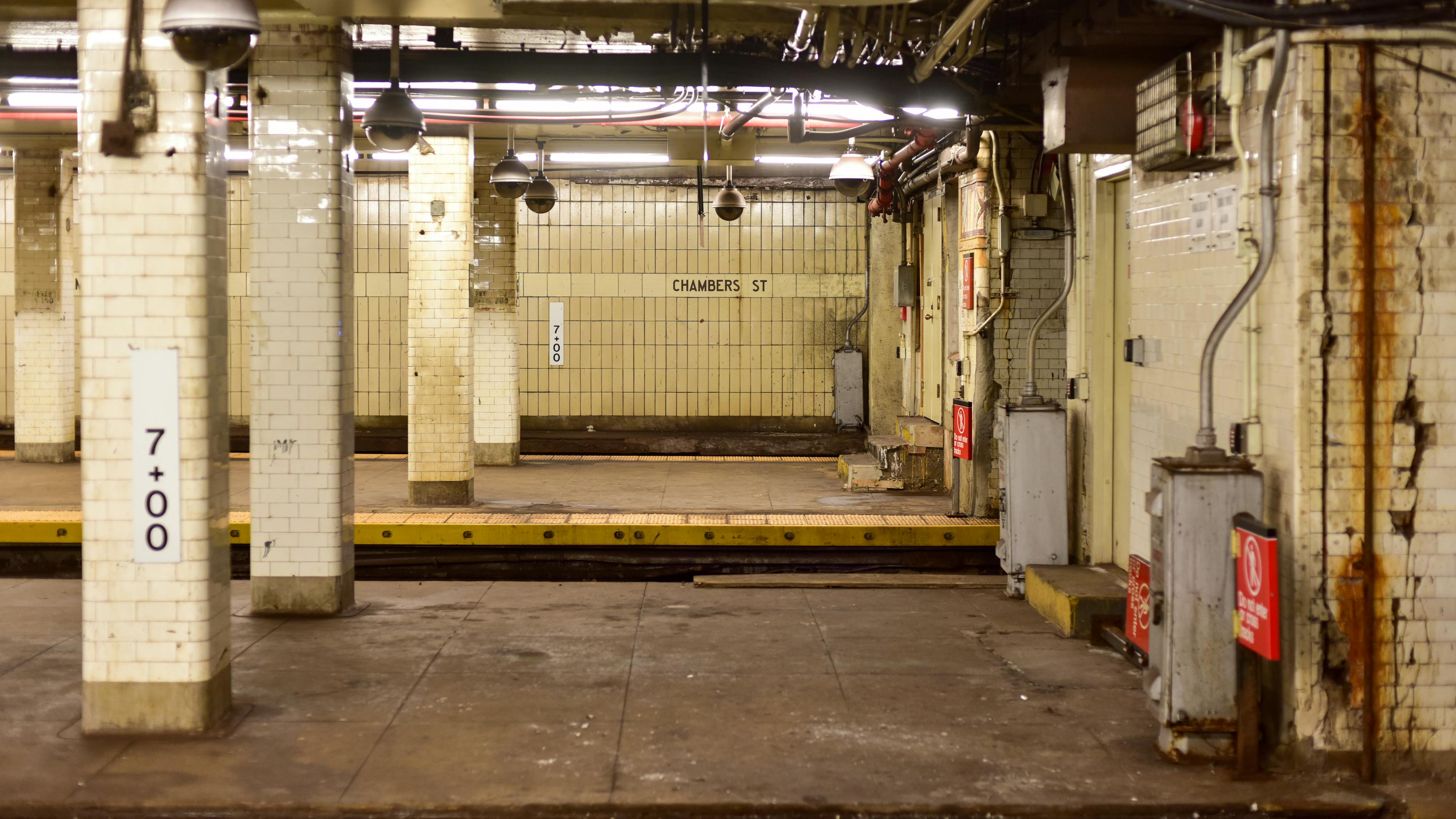 Empty New York City subway platform at the Chambers Street station, with white tile columns, a station wall marked 'Chambers St,' ceiling-mounted security cameras, and visible wear and rust along the walls.