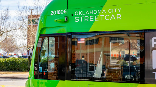Close-up of an Oklahoma City Streetcar vehicle numbered 201806, showing its green livery, forward-facing security camera, and front windshield with an American flag decal, parked with bare trees and a building in the background.
