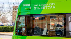 Close-up of an Oklahoma City Streetcar vehicle numbered 201806, showing its green livery, forward-facing security camera, and front windshield with an American flag decal, parked with bare trees and a building in the background.