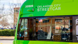 Close-up of an Oklahoma City Streetcar vehicle numbered 201806, showing its green livery, forward-facing security camera, and front windshield with an American flag decal, parked with bare trees and a building in the background.