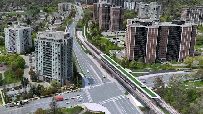 An aerial rendering of the elevated guideway section of the Eglinton Crosstown West Extension in Toronto, Ontario, showing a light-rail transit structure running alongside a multi-lane arterial road through a residential neighborhood of mid-rise and high-rise apartment towers, with green landscaping along the guideway base and a station platform visible in the foreground.