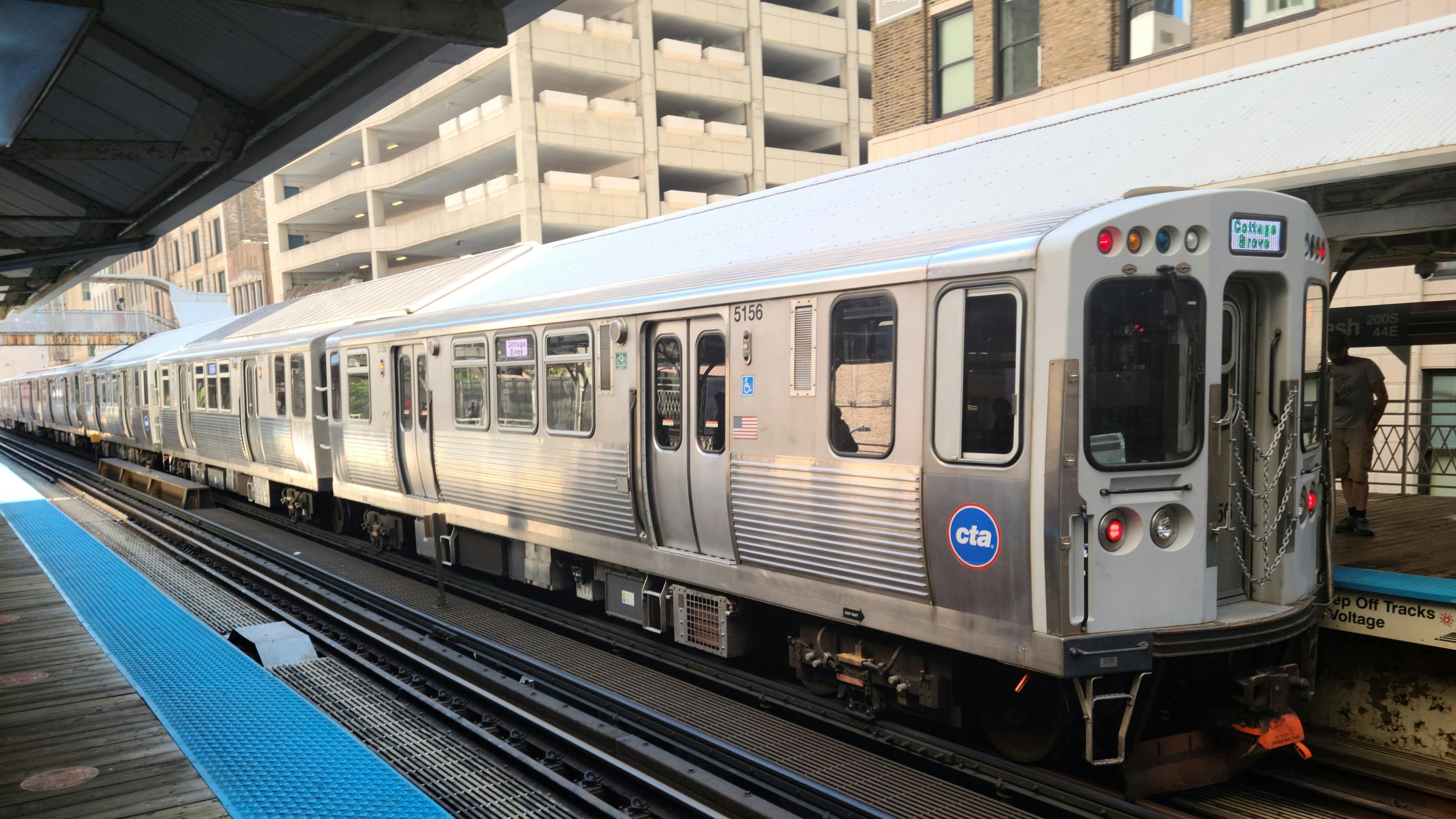 A CTA 'L' train, car 5156, displaying a 'Cottage Grove' destination sign, stopped at an elevated Chicago platform with the agency's blue circular logo on the front car, mid-rise buildings visible in the background.