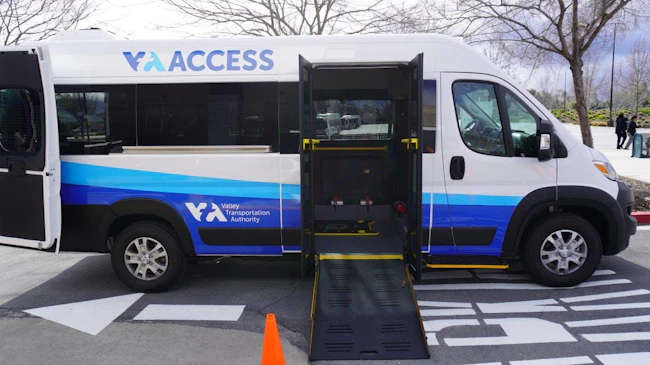 A Santa Clara Valley Transportation Authority ACCESS Paratransit van in white and blue livery with its side door open and ADA wheelchair ramp deployed, parked in a transit facility lot with an orange safety cone placed at the base of the ramp.
